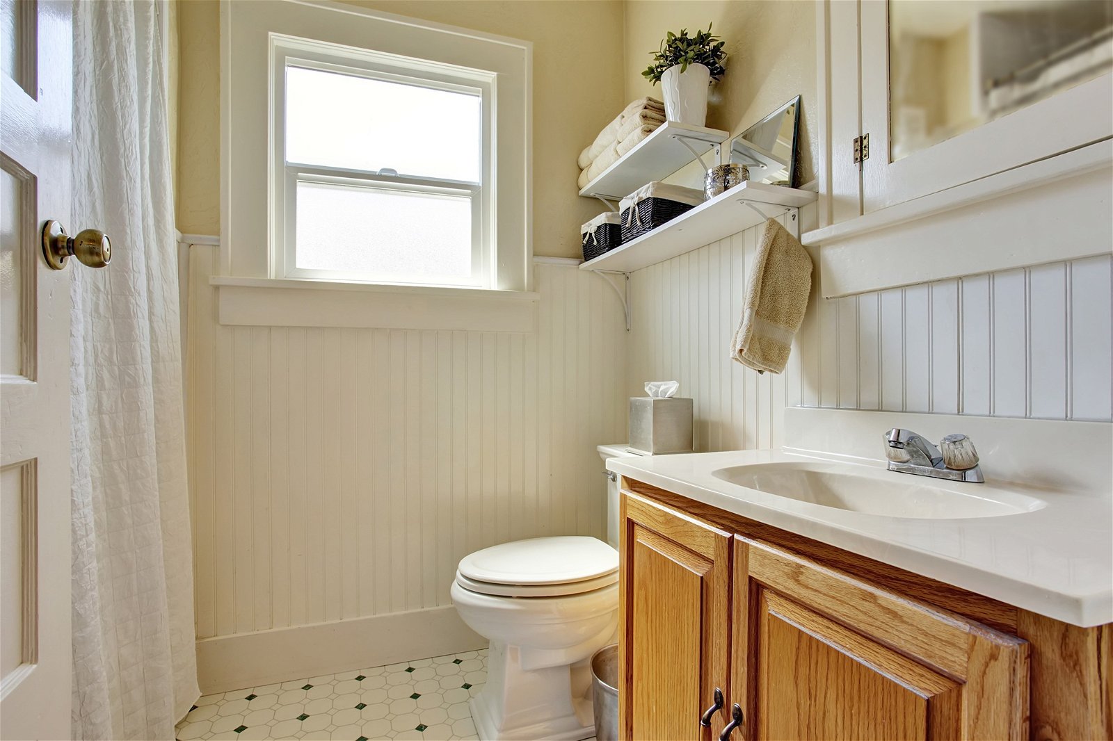 Bathroom design in creamy colors with brown wooden cabinet and small window. Decorated with plant pot on the shelf.