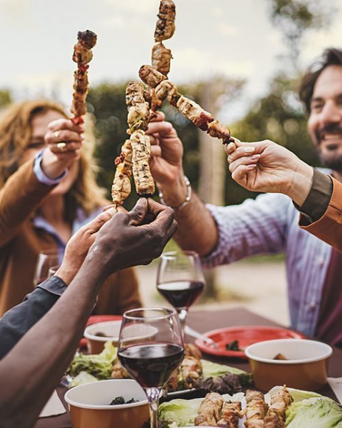 Toasting with Skewers - Group of people in their 30s and 40s at a garden table, holding meat skewers and toasting. Mixed ethnicity. Blurred trees and sunset in the background.