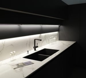Modern kitchen with black furniture, White marble worktop and backsplash.Black sink and tap, Light under wall cabinet.